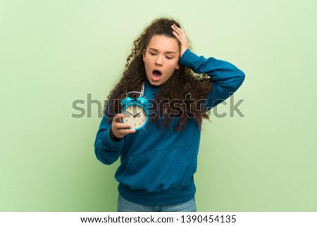 Teenager girl over green wall restless because it has become late and holding vintage alarm clock