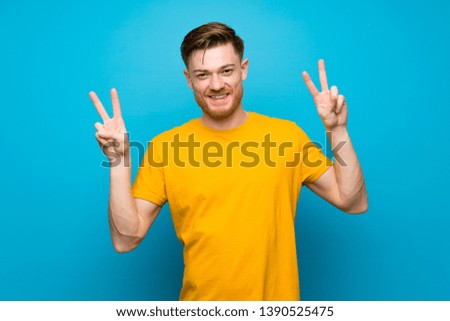 Redhead man over blue wall showing victory sign with both hands