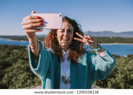 A young brunette woman is taking a picture of herself in the middle of the nature. She is sticking out tongue and making the symbol of victory with his other hand at the camera.