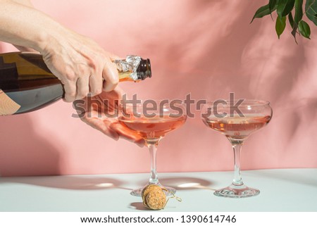 Female hand pouring champagne or wine into glasses. Gentle pink background bright sunlight. Copy space minimalism.
