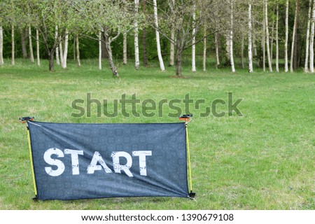 Start - word on grey fabric banner for orienteering or rogaining sport outdoors on green grass field and tree trunks background with copy space.
