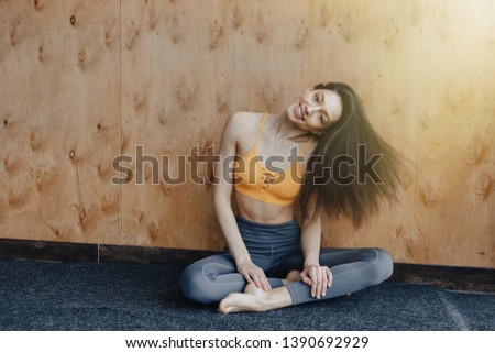 Young attractive fitness girl sitting on the floor near the window on the background of a wooden wall, resting on yoga classes at gym