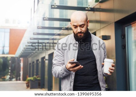 Portrait of young smiling man using smartphone on city street,bearded hipster guy communicates with friends in social networks via mobile phone outdoors.Man sends text message,drinks coffee.Lifestyle.