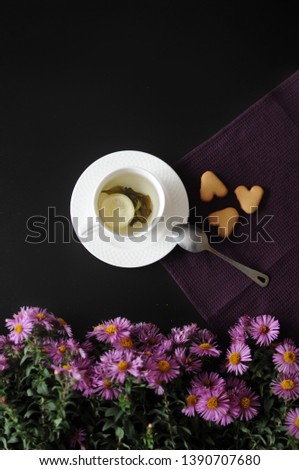 White cup of green tea and lemon on lace napkin with homemade hurt cookies, spoon, purple flowers on background is on black table