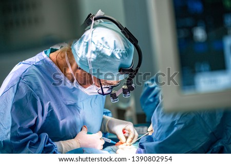 Surgeon and his assistant performing cosmetic surgery in hospital operating room. Surgeon in mask wearing loupes during medical procadure. Royalty-Free Stock Photo #1390802954