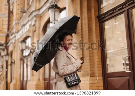 Beautiful woman in beige trench coat with cross-body bag with smile walks under umbrella in European city