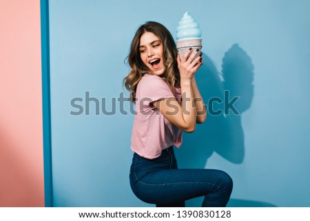 Fashionable woman in jeans dancing on blue background. Glad brunette girl eating ice cream.