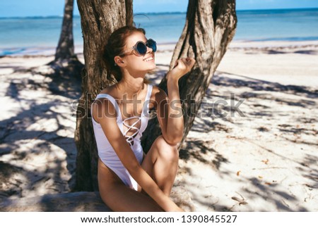 A tanned woman in a white bathing suit and wearing glasses sits near a palm tree on an island             