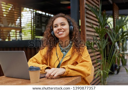 Photo of young dark skinned curly student lady siting on a cafe terrace, listens to music and dreamily looking away, wearing in yellow coat, drinking coffee, works at a laptop.