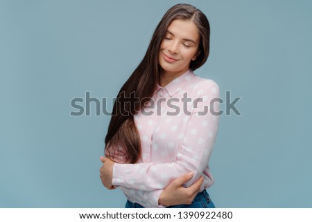 Photo of pleased woman enjoys daydreaming, embraces herself, keeps eyes closed, tilts head, has long thick dark hair, wears fashionable clothing, models over blue background, expresses happiness