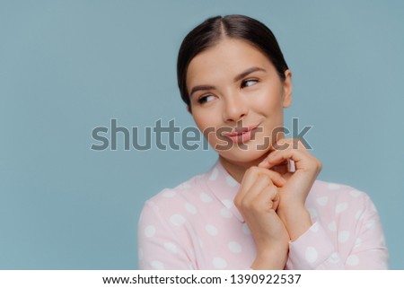 Close up shot of dreamy beautiful lady keeps hands together, focused aside, had black hair, healthy skin, dressed in polka dot shirt, poses over blue background with empty space for your promotion