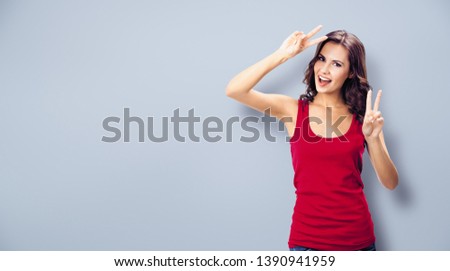 Portrait photo - young smiling beautiful woman in casual clothing, showing two fingers or victory gesture, on grey wall background. Happy girl in red dress. Brunette excited model at studio picture.