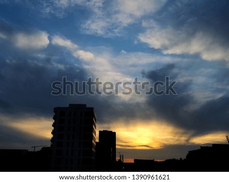 Photo of a city skyline as seen at sunset, with 2 tall buildings standing out to the right of the picture. The sky is yellow, grey, and blue due to heavy clouds.