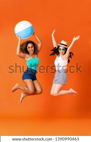 Happy African American and Asian woman friends in casual summer clothes with colorful beach ball jumping in coral orange studio background