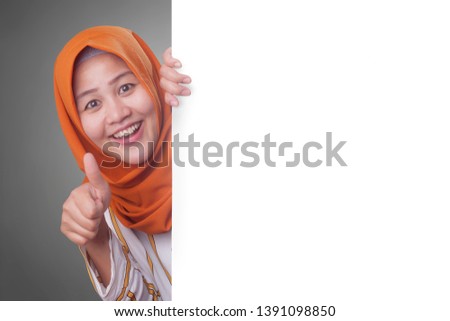 Young Asian muslim businesswoman smiling and peeking behind blank white advertising billboard wign board with copy space
