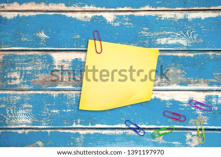 One blank square paper sticker with red staple near lot of scattered clips on worn blue wooden table. Top view. Back to school or business concept. Copy space