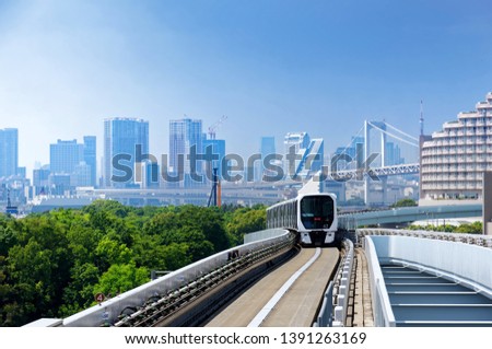A metro train traveling on the elevated track of Yurikamome Line in Odaiba, with the landmark Rainbow Bridge crossing Tokyo Bay & modern skyscrapers clustering in Minato District under blue sunny sky Royalty-Free Stock Photo #1391263169