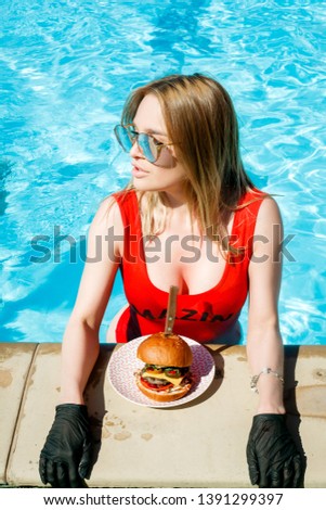 Burger, stands against the background of a large breast of a girl, in a red swimsuit, in the pool. The concept of fast food on vacation