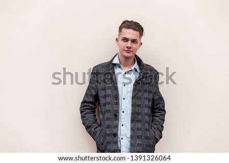 Young attractive guy with a stylish hairstyle in a long gray checkered jacket in a fashionable shirt is standing near a white wall on the street on a spring day. Modern nice guy is enjoying a walk.