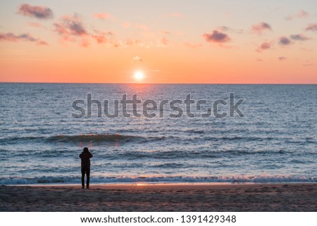 Very beautiful sunset on the Baltic Sea. In the foreground a young man is photographing the illuminated splashing of the waves.
The sea is calm.