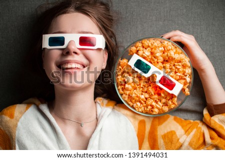 young cute girl lies on a gray background in 3d glasses eats popcorn and watches a movie in her pajamas