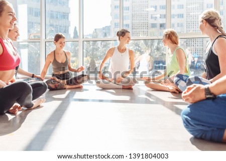 Group of young positive women are doing padmasana sitting on the floor in circle during group classes in a spacious hall on the background of large windows. Concept of women's communication and health