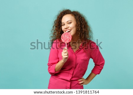 Portrait of cute african girl in casual clothes hold in hand eating pink round lollipop isolated on blue turquoise background in studio. People sincere emotions, lifestyle concept. Mock up copy space