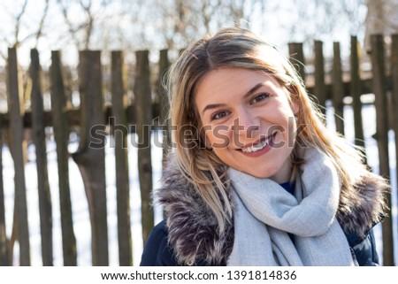 Portrait of a beautiful young woman wearing warm coat and grey scarf , smiling to the camera in front of vintage fence