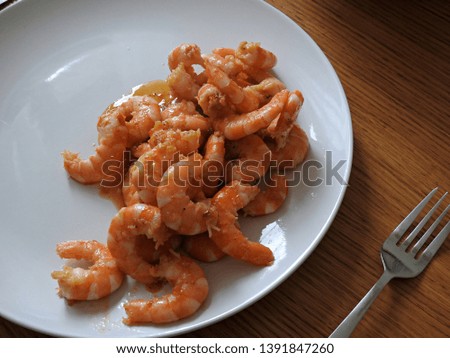 Shrimp/prawn cooked in garlic served on a round white plate on a wooden table with fork, diagonal top view. Minimalistic serving of seafood dish