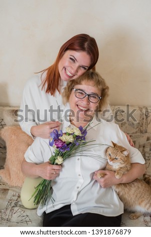 Daughter gives a bouquet to an adult mother sitting on a sofa in the living room. Spending time together, celebrating at home on weekends. Mothers Day. Cat eats flowers