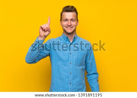 Blonde man over isolated yellow wall showing and lifting a finger in sign of the best
