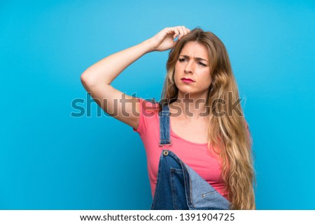 Young blonde woman with overalls over isolated blue wall having doubts while scratching head