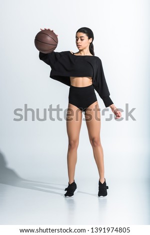 attractive sportive african american girl in black sportswear and sneakers playing ball on white