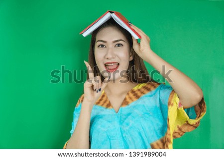 Pensive attractive asian girl holds book on head and looks, has straight hair,  with school object, holds textbooks, she has interesting poses against green background. Studying concept.