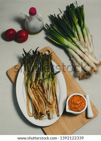 Overhead picture of baked vegetables called calcots on wooden board accompanied by sauce and ingredients such as tomato and oil