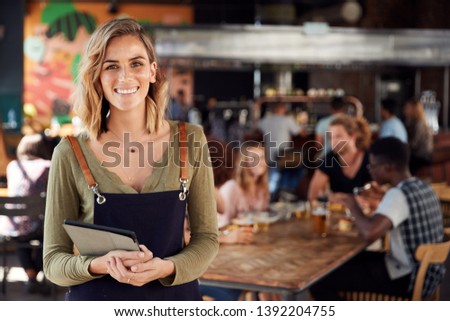 Portrait Of Waitress Holding Menus Serving In Busy Bar Restaurant Royalty-Free Stock Photo #1392204755