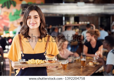 Portrait Of Waitress Serving Food To Customers In Busy Bar Restaurant Royalty-Free Stock Photo #1392204758