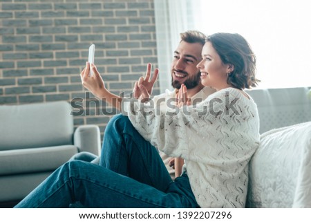 happy woman waving hand while taking selfie with man showing peace sign at home 