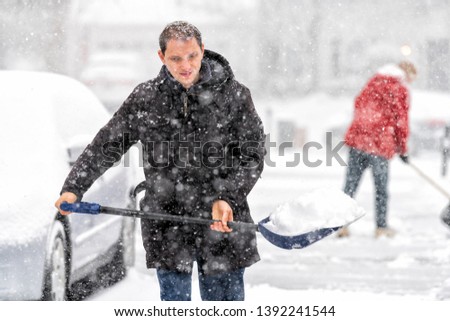 Happy young man in winter coat cleaning shoveling driveway street from covered falling snow in heavy snowstorm with shovel