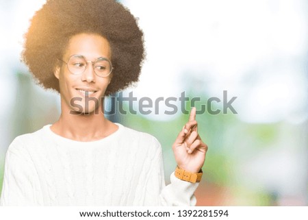 Young african american man with afro hair wearing glasses with a big smile on face, pointing with hand and finger to the side looking at the camera.
