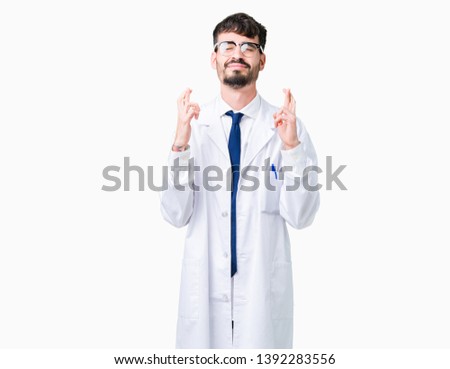 Young professional scientist man wearing white coat over isolated background smiling crossing fingers with hope and eyes closed. Luck and superstitious concept.