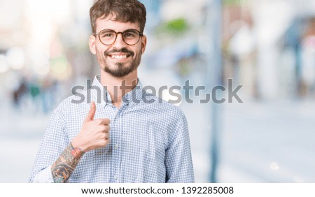Young handsome man wearing glasses over isolated background doing happy thumbs up gesture with hand. Approving expression looking at the camera with showing success.