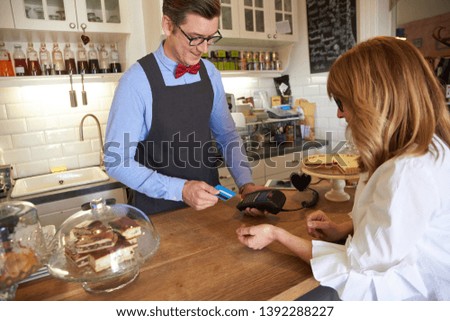 Shot of young waitress standing behind the counter in small cafe while her guest paying with bank card. Small business. 