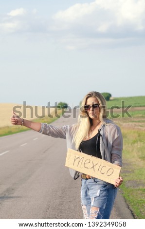 Blonde holding sign while hitchhiking on the road in summertime.