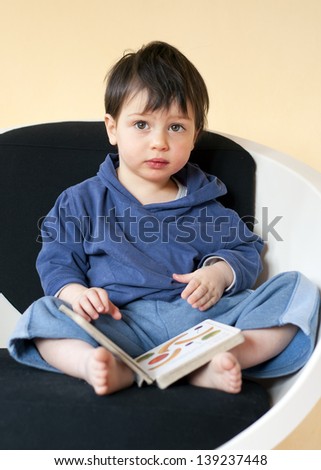Toddler child sitting on a  chair at home reading a book.
