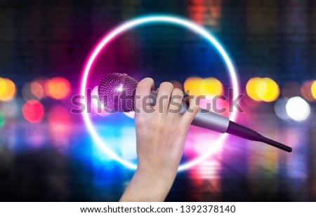 A female hand is holding one microphone against the colorful lights of the karaoke club scene. Bright colorful background with blurred neon lights
