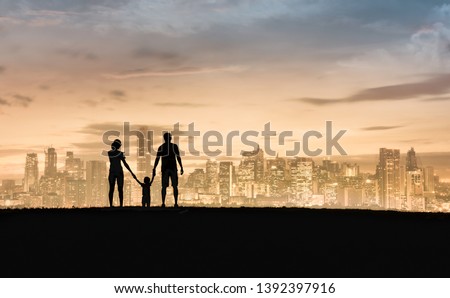 Family of three silhouette father, mother, child standing on a hill looking at city view holding hands. 