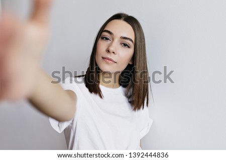 Good-looking european woman making selfie on white background. Inspired young lady with dark hair taking picture of herself with serious face expression.