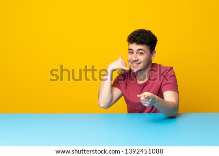 Young man with colorful wall and table making phone gesture and pointing front