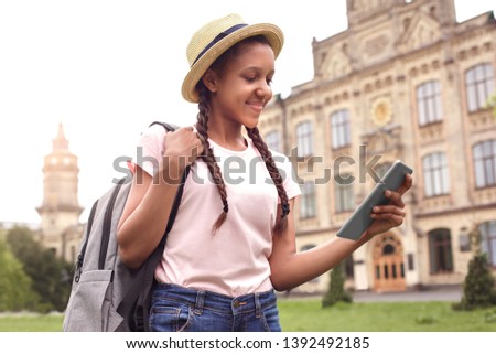 Young african american girl wearing hat at school yard walking carrying backpack holding digital tablet watching cartoon online concentrated smiling happy